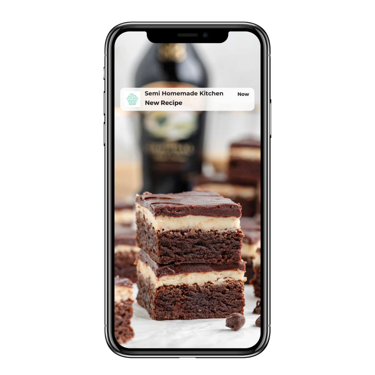 Image of a smartphone displaying a close-up of chocolate and cream layered brownies. A notification on the screen reads Semi Homemade Kitchen New Recipe. A bottle is blurred in the background.