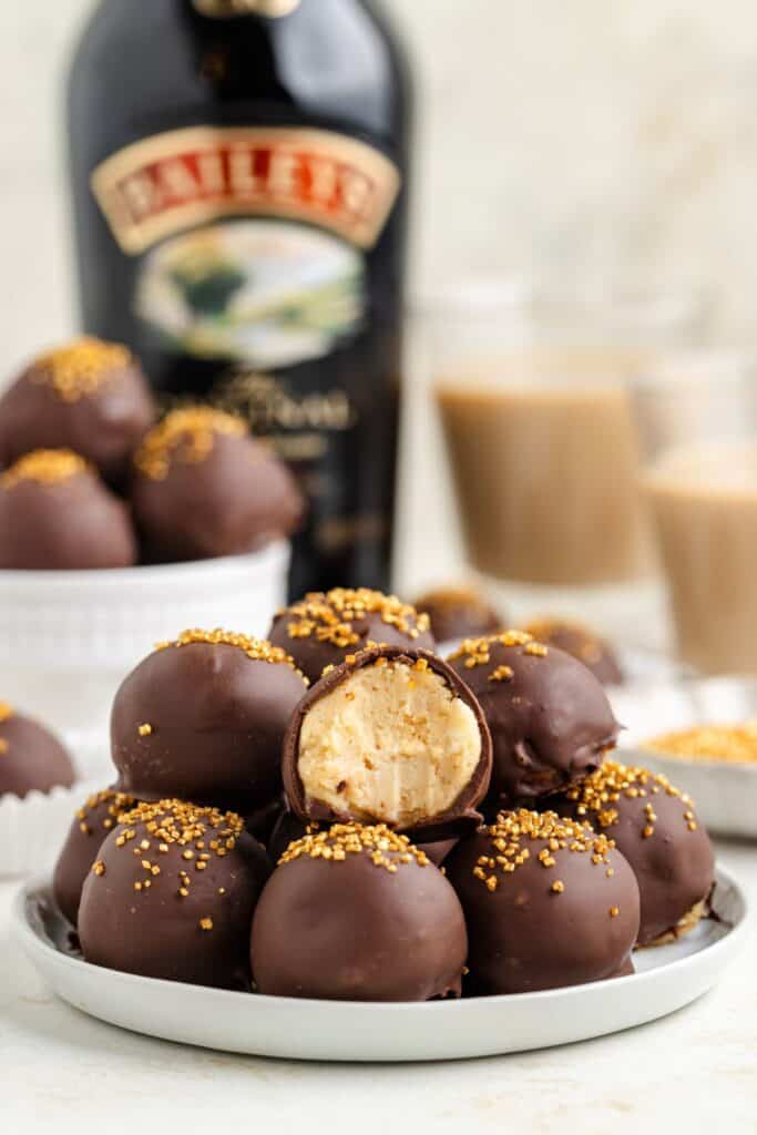 A plate of chocolate-covered Baileys truffles with golden sprinkles, one is halved showing a creamy center. A bottle of Baileys and glasses of creamy drinks are blurred in the background.