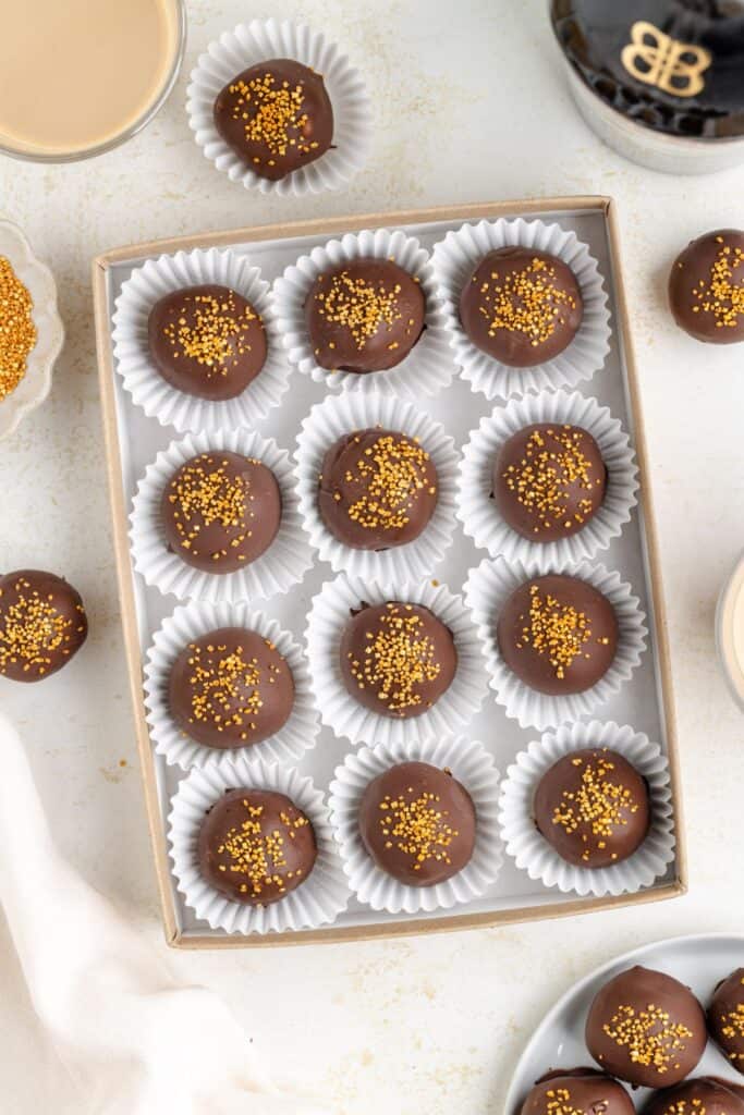 A box of Baileys truffles placed in white paper cups, each topped with golden sprinkles. Nearby, there are glasses with a Baileys, a few extra truffles, and a small bowl with golden sprinkles on a light surface.