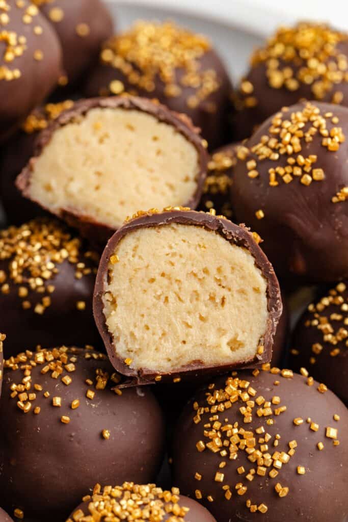 Close-up of chocolate-covered Baileys truffles with golden sprinkles. One truffle is cut in half, revealing a creamy, light brown filling inside. The rest are whole and coated with chocolates and sprinkles, placed on a plate.