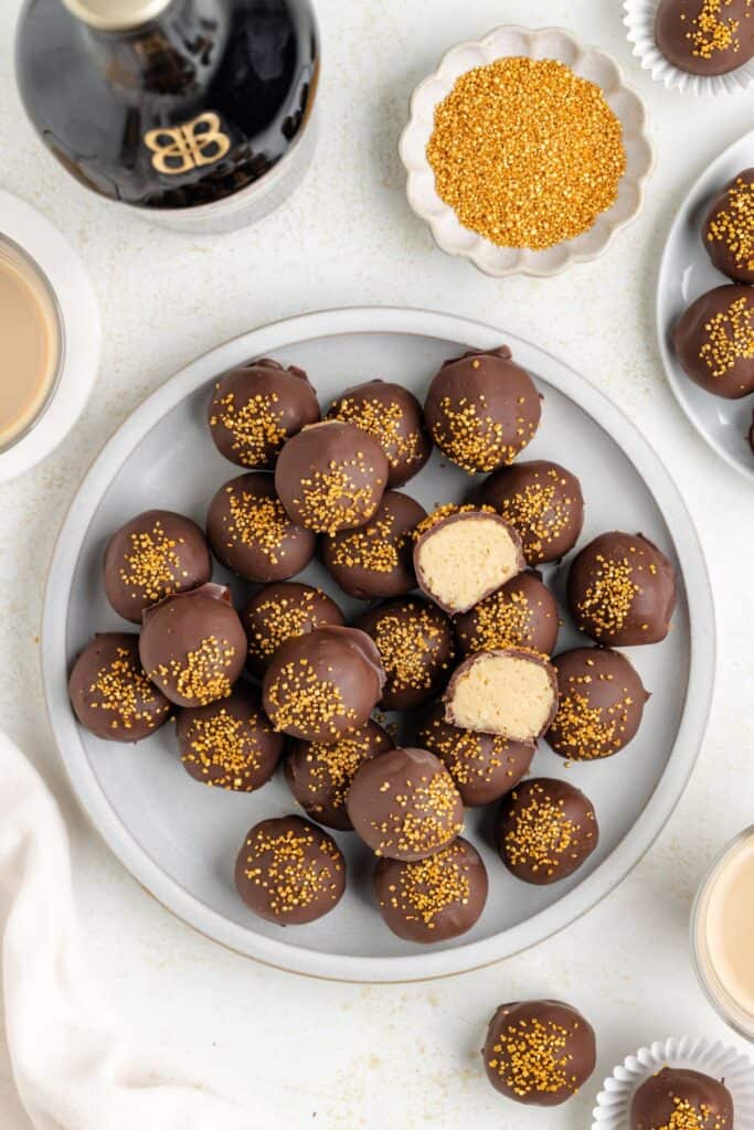 A gray plate filled with chocolate-covered Baileys balls sprinkled with golden decorations. One ball is cut in half, revealing a creamy filling. Nearby are small dishes with golden sprinkles and drinks, along with a bottle of Baileys.