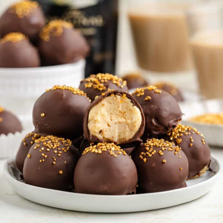 A plate stacked with chocolate-covered Baileys truffles, some topped with golden sprinkles. One truffle is cut in half, revealing a creamy filling. More truffles and drinks are blurred in the background.