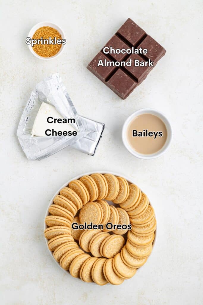 Top view of ingredients on a white surface. There are golden Oreos on a round plate, an unwrapped block of cream cheese, a bowl of sprinkles, squares of chocolate almond bark, and a cup of Baileys. Labels identify each item.