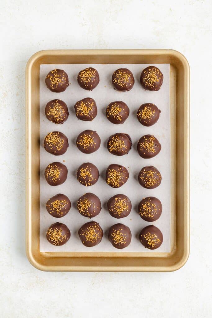 A gold baking tray holds 24 uniformly arranged chocolate-covered Baileys balls, each topped with golden sprinkles. The tray sits on a light-colored surface.