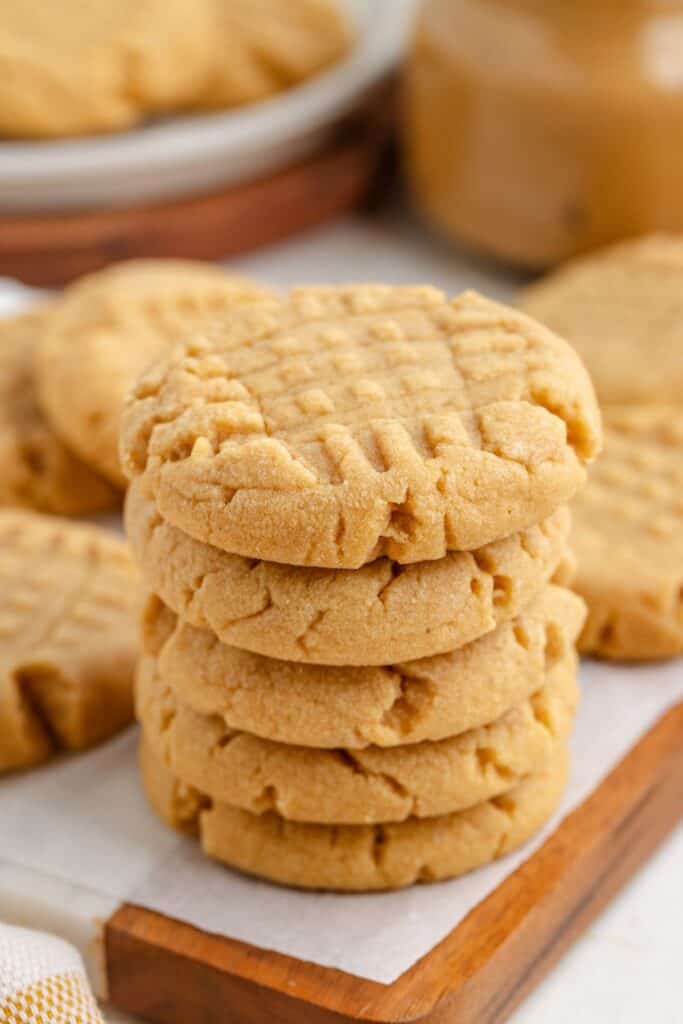 A stack of five cake mix peanut butter cookies with a crisscross pattern on top, placed on a wooden board. More cookies and a jar of peanut butter are blurred in the background.