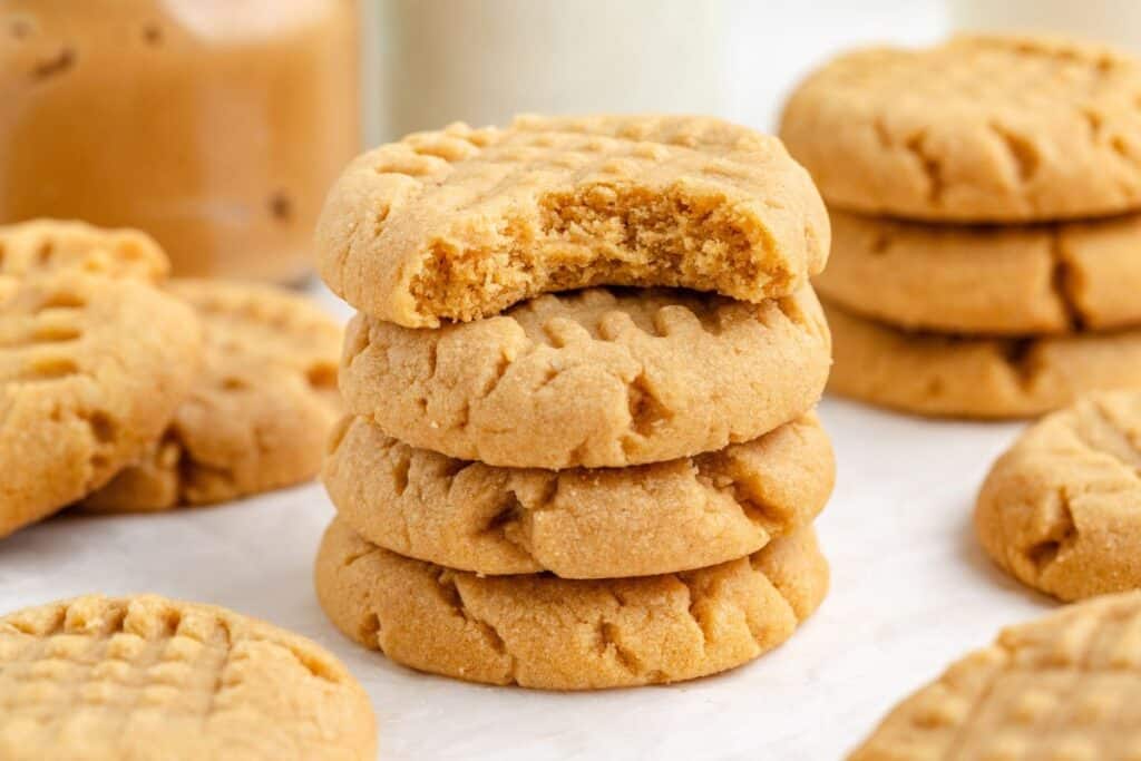 A stack of four peanut butter cookies, with the top one partially eaten, sits on a light surface. More cookies and a jar of peanut butter are blurred in the background. The cookies have a crisscross pattern on top.