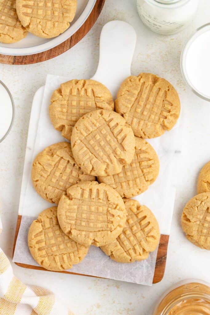 A wooden board holds a pile of peanut butter cookies made with cake mix with crisscross fork patterns. A glass of milk and a jar are nearby, along with additional cookies on the side.