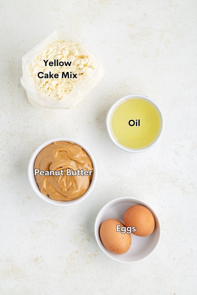 Labeled ingredients for a recipe are displayed on a light surface: a bag of yellow cake mix, a bowl of peanut butter, a bowl of oil, and two eggs.