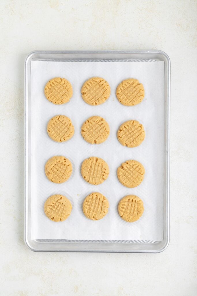 A baking tray with twelve cake mix peanut butter cookies placed on white parchment paper. The cookies have a crisscross pattern on top. The tray is set against a light background.