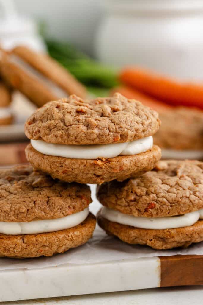 Three carrot cake sandwich cookies with cream cheese filling, stacked on a marble surface, surrounded by more cookies and carrots in the background.