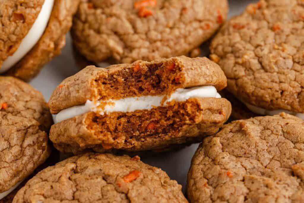 Close-up of carrot cake cookies with cream cheese filling, one of which is bitten, revealing its texture and carrot bits.