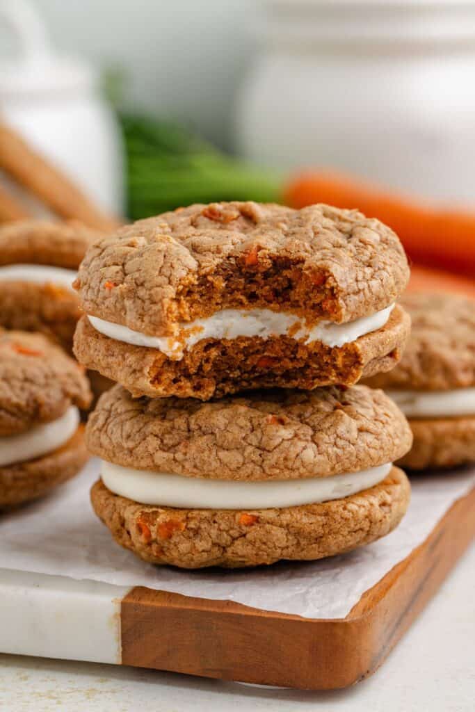 Two sandwich-style carrot cookies with cream cheese filling on a wooden tray; one cookie has a bite taken out.