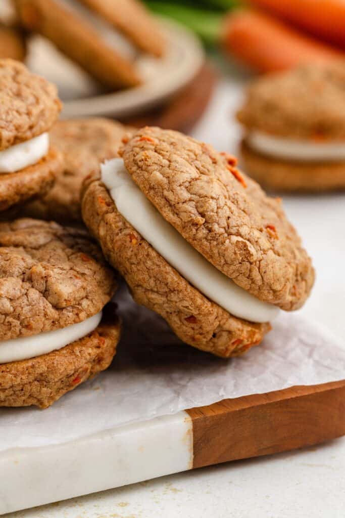 Close-up of carrot cake sandwich cookies filled with cream cheese frosting, stacked on parchment paper over a marble and wood surface.