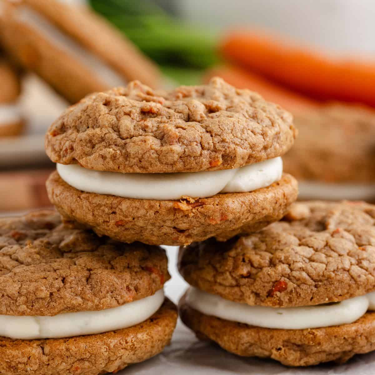 Three carrot cake cookies with cream cheese filling stacked on a surface, surrounded by blurred carrots in the background.