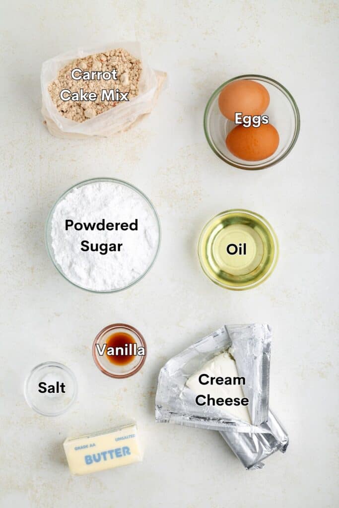 Ingredients for a carrot cake mix recipe arranged on a white surface: carrot cake mix, eggs, powdered sugar, oil, vanilla, salt, butter, and cream cheese.