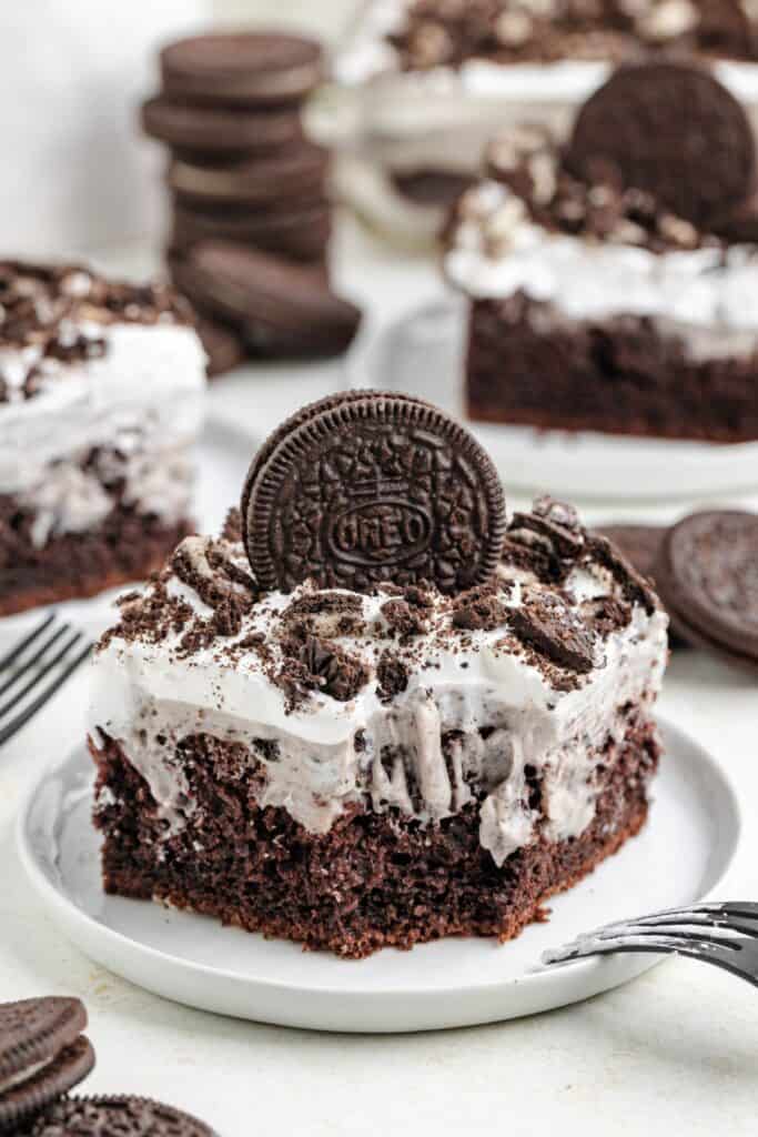 A square piece of Oreo Poke Cake with Cool Whip topped with crumbled cookies and a whole cookie sits on a plate, surrounded by additional cake slices and stacked cookies in the background.