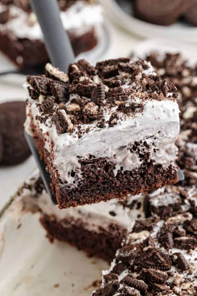 A spatula lifts a slice of Oreo Poke cake topped with whipped topping and crushed Oreo cookies.