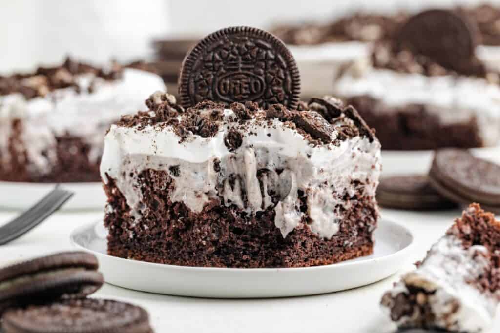 A chocolate poke cake infused with Oreo pudding, topped with crumbled cookies and cream, and garnished with a whole cookie on a white plate.