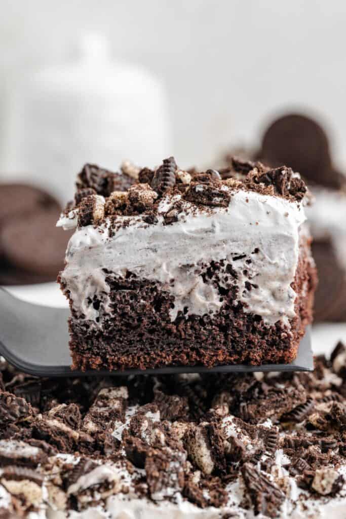 A slice of Oreo pudding poke cake topped with white cream and crumbled Oreo cookies on a spatula.