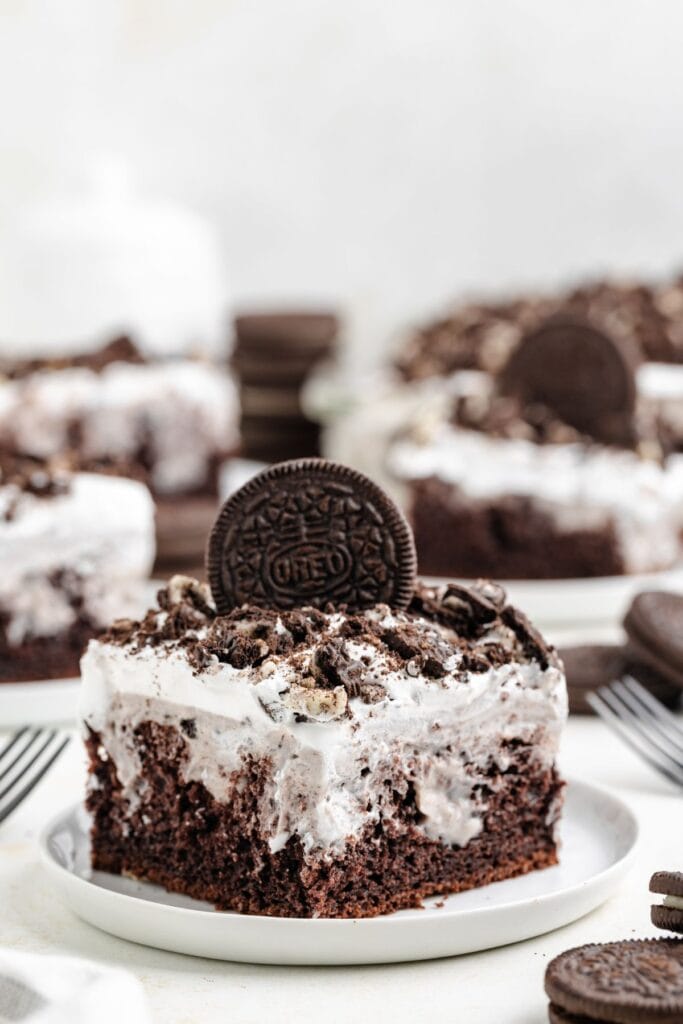 A slice of chocolate Oreo poke cake topped with whipped topping, crumbled Oreo cookies, and a whole cookie on a white plate.