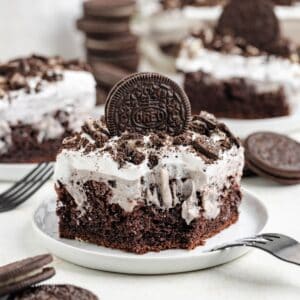 A slice of Oreo poke cake with Cool Whip frosting, topped with crushed Oreos and a whole cookie, served on a white plate.