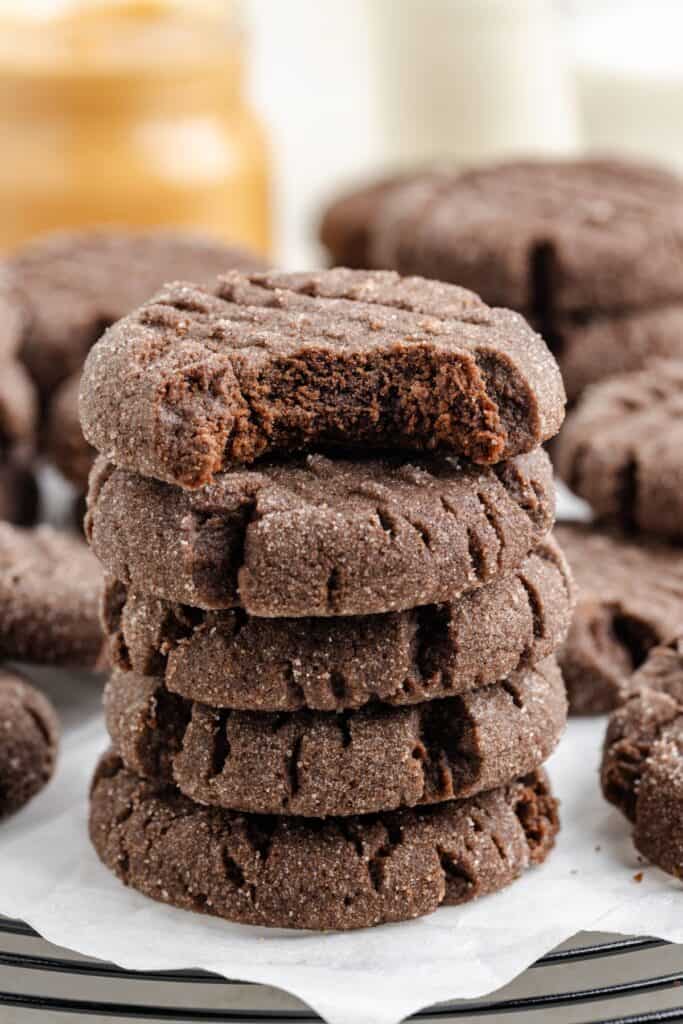 A stack of five sugar-coated peanut butter chocolate cake mix cookies is on a tray, with a bite taken out of the top cookie. Other cookies are scattered in the background, with blurred jars and glasses in the back.