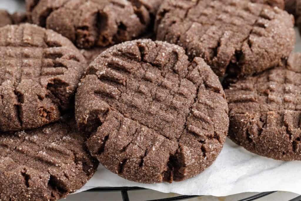 Close-up of several chocolate peanut butter cake mix cookies with a crisscross pattern on top, lightly coated with sugar. They are arranged in a slightly overlapping manner on white parchment paper.