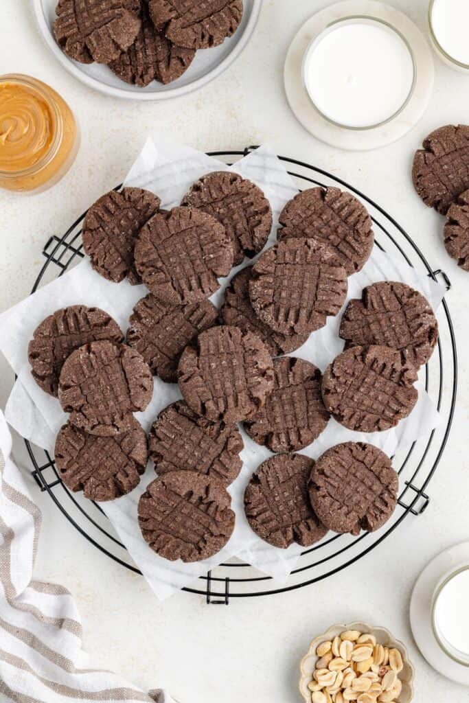 Peanut butter chocolate cake mix cookies with a crisscross pattern on top, placed on white parchment paper on a circular cooling rack. Surrounding items include a jar of peanut butter, a small bowl of peanuts, glasses of milk, and a striped cloth.