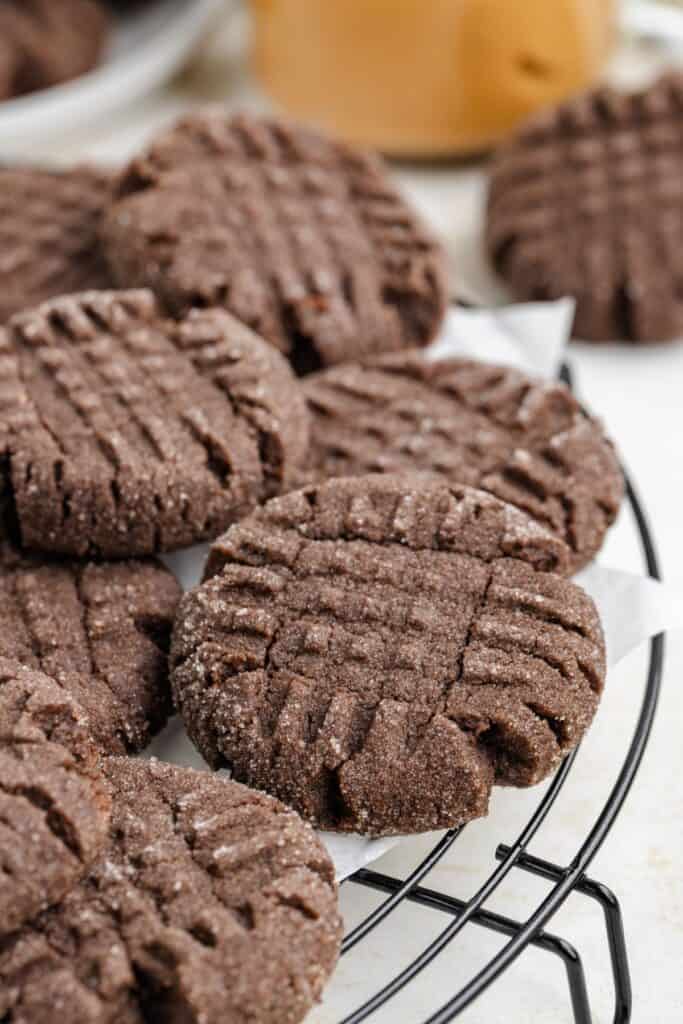 Close-up of freshly baked chocolate peanut butter cookies with a crisscross pattern, dusted with sugar, cooling on a black wire rack. A few more cookies are visible in the blurred background.