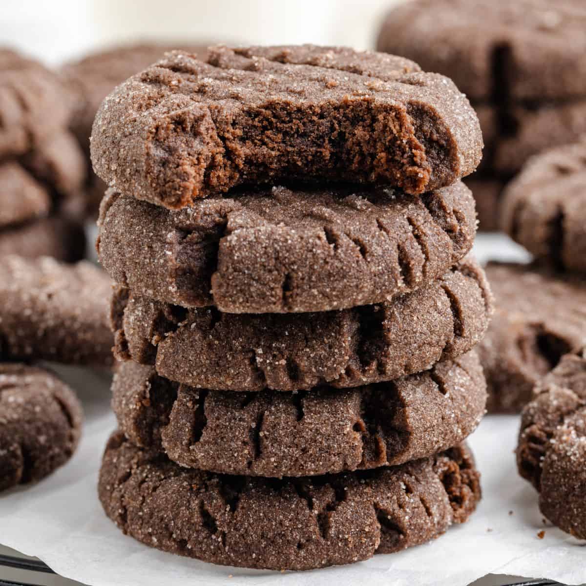 A stack of five chocolate peanut butter cake mix cookies is shown, with the top cookie having a bite taken out of it. The cookies are dark brown and textured, resting on parchment paper with more cookies blurred in the background.