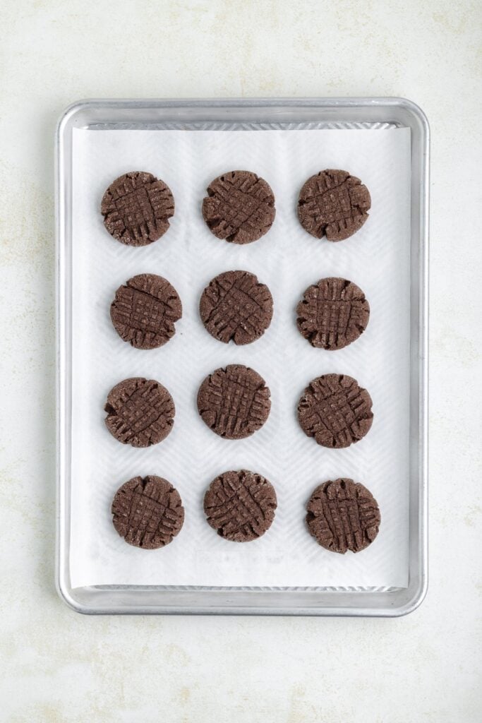 A baking tray with twelve evenly spaced round chocolate peanut butter cookies on parchment paper. Each cookie has a crisscross pattern on top. The tray is placed on a light-colored surface.