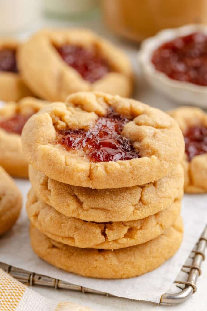 A stack of peanut butter and jelly cake mix cookies on a wire rack, with more cookies and a bowl of jelly in the background.