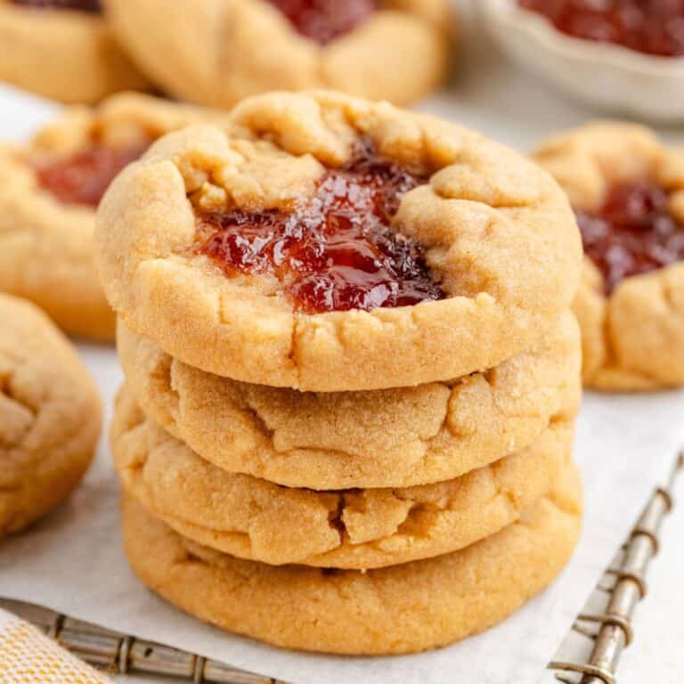 A stack of four peanut butter and jelly cake mix cookies on a wire rack, with more cookies and a bowl of jelly in the background.