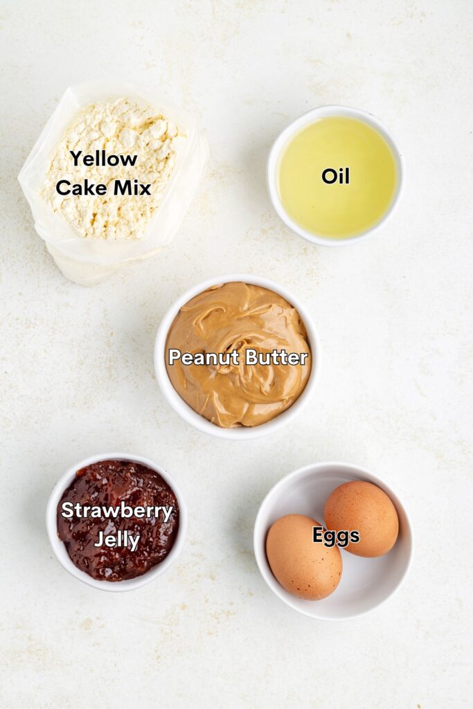 Ingredients for baking arranged: yellow cake mix in a bag, a bowl of oil, a bowl of peanut butter, a bowl of strawberry jelly, and two eggs in a bowl.