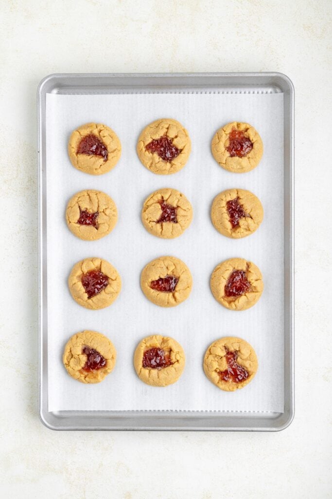 A baking tray with twelve peanut butter cookies, each topped with a dollop of red jam, arranged in rows on parchment paper.