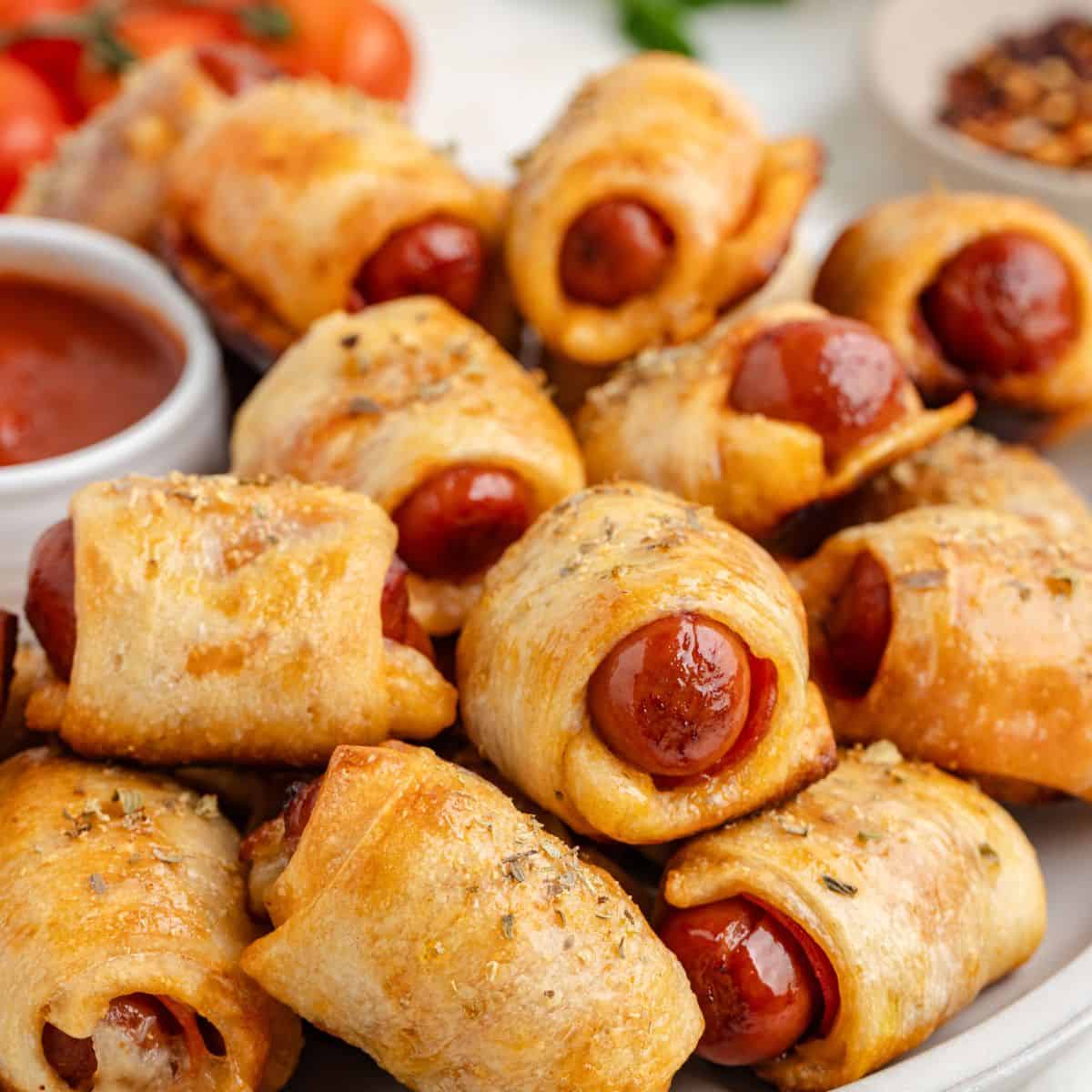 Close-up of a plate filled with baked pizza pigs in a blanket. There are bowls of marinara sauce and chili flakes in the background, along with cherry tomatoes.