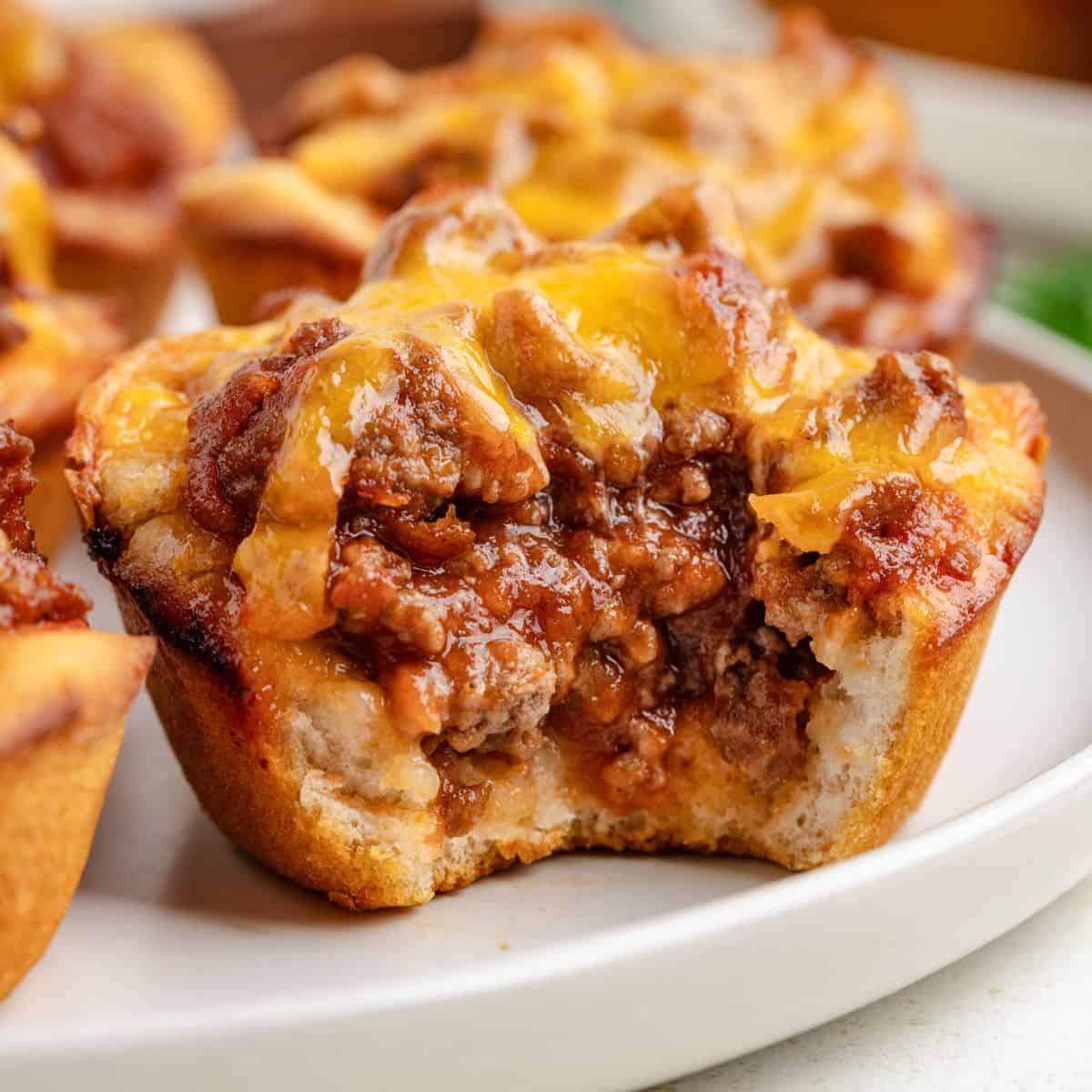 Close-up of a Sloppy Joe Biscuit Cup filled with ground meat and melted cheese, served on a white plate. A bite is taken from the front.