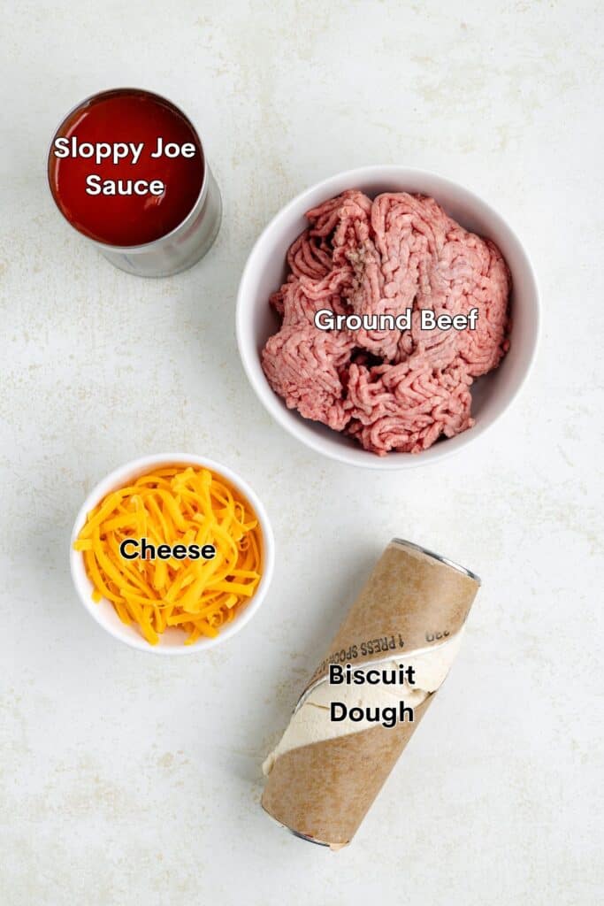 Ingredients laid out: a can of Sloppy Joe sauce, a bowl of ground beef, a bowl of shredded cheese, and a tube of biscuit dough on a light surface.