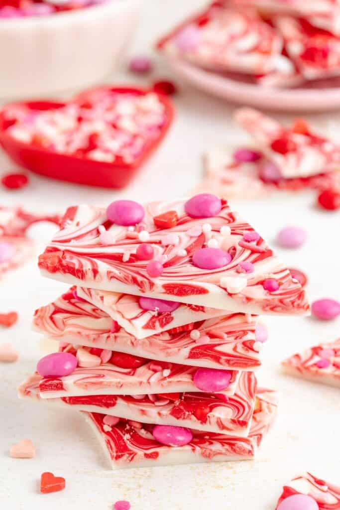 Stack of red and white swirled candy bark with pink and red sprinkles, surrounded by heart-shaped bowls filled with similar treats.