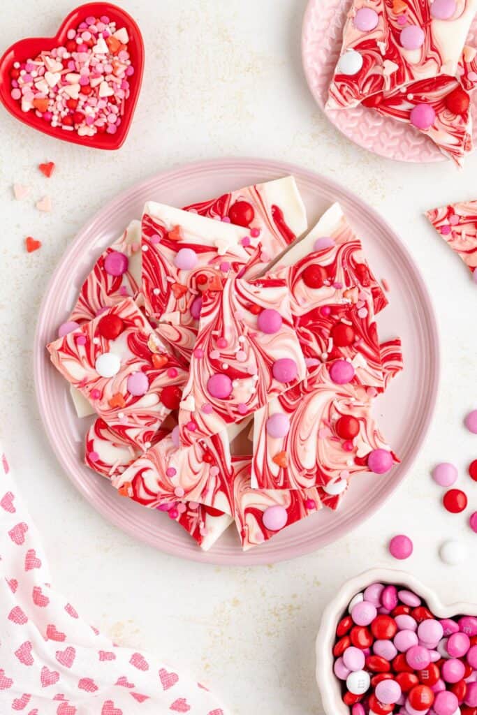 Plate of swirled pink, red, and white chocolate bark topped with colorful candies, surrounded by heart-shaped sprinkles and candy dishes.