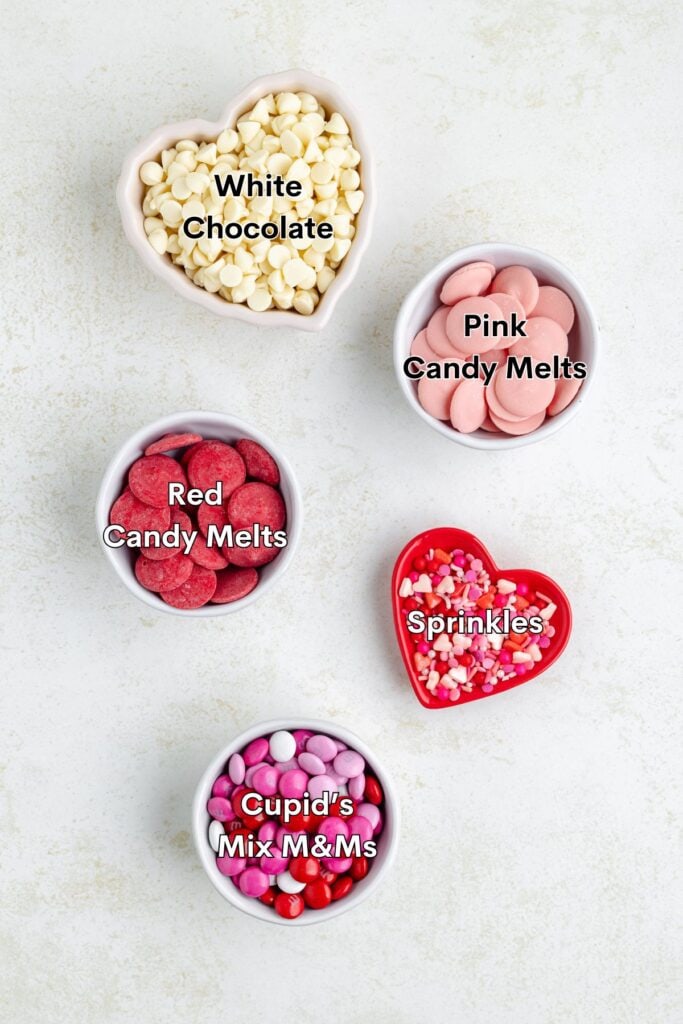 Five bowls containing white chocolate chips, pink candy melts, red candy melts, heart sprinkles, and Cupids Mix M&Ms on a white surface.