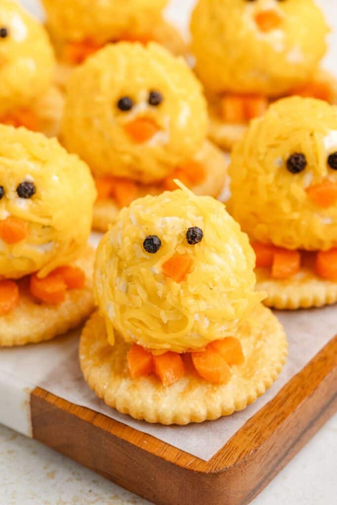 Mini cheese ball chicks on crackers, adorned with eyes and beaks, on a wooden tray. Festive setting.