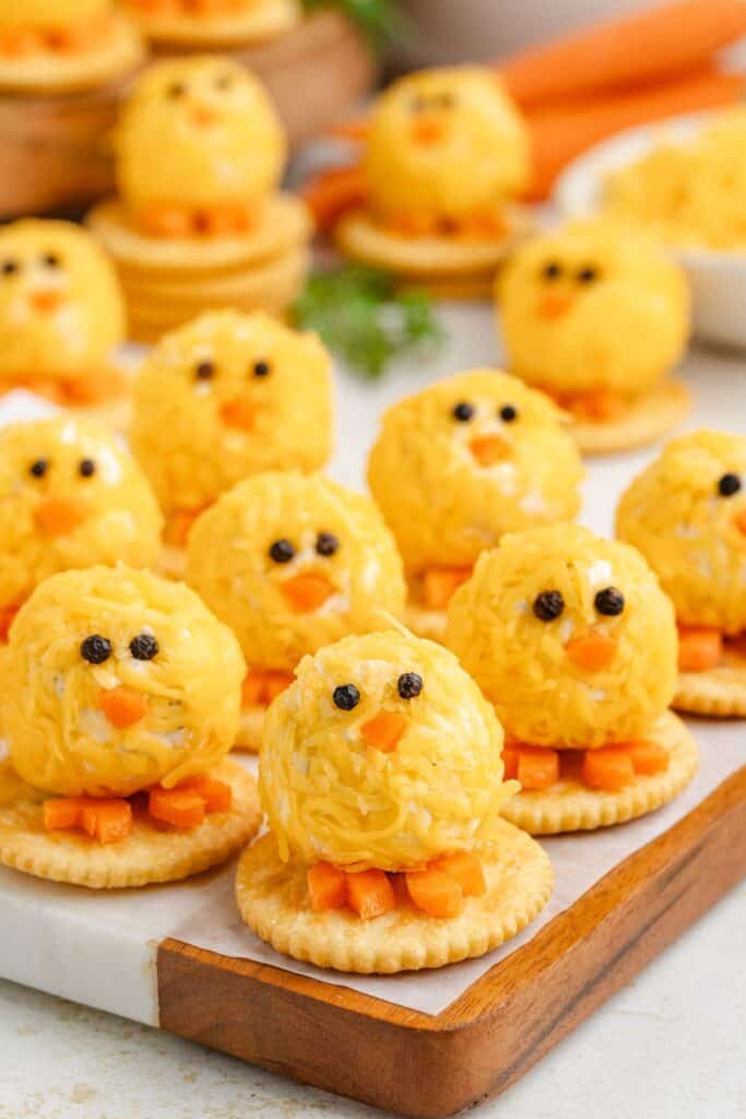 Cheese balls shaped like chicks on crackers, with carrot beaks, arranged on a wooden tray.