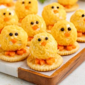 Baby chicks cheese ball with peppercorn eyes, carrot beaks and feet, on crackers on a wooden tray.