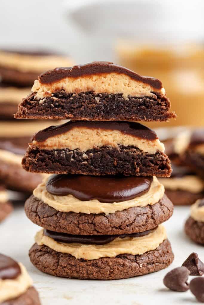 A stack of Buckeye Brownie Cookies with creamy peanut butter layers, topped with chocolate glaze. Two cookies on top, one halved, showcasing their filling.