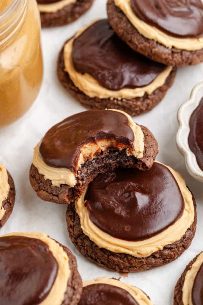 Chocolate cookies with peanut butter frosting and chocolate glaze. One cookie has a bite taken out of it. A jar of peanut butter is visible in the background.
