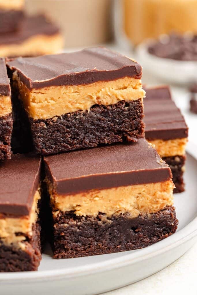 Three-layer buckeye brownies stacked on a plate. Bottom layer is brownie, middle is peanut butter, and top is chocolate.