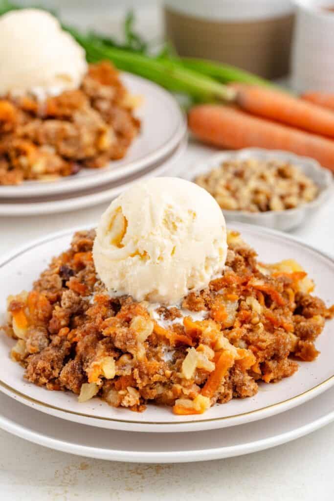 A plate of carrot dump cake topped with a scoop of vanilla ice cream. Carrots and chopped nuts are in the background.