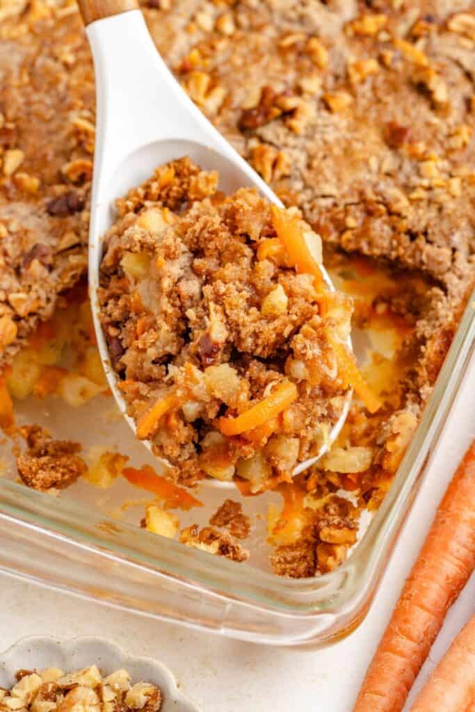 A spoon holds a scoop of baked carrot and pineapple dump cake in a glass dish surrounded by pieces of carrot.