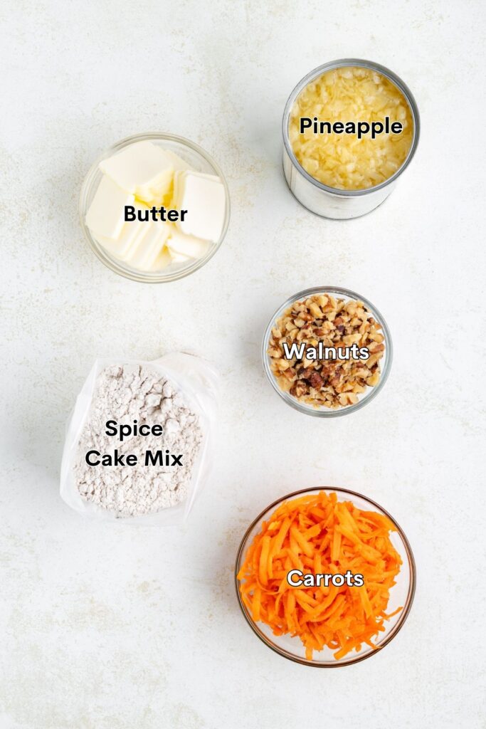 Ingredients for a recipe are laid out on a surface: butter, pineapple, walnuts, spice cake mix, and grated carrots.